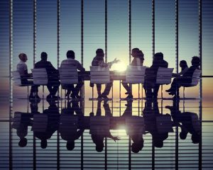 Image is of a business meeting taking place Infront of a glass window where the sun is rising in the background.